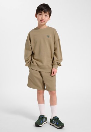 Jongen draagt een oversized beige sweatshirt en bijpassende korte broek, met witte sokken en donkergroene sneakers, staand tegen een witte achtergrond.