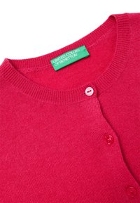 Fuchsiový kardigan vyrobený z pletené látky, s kulatým výstřihem, dvěma červenými knoflíky a zelenou cedulkou s nápisem "UNITED COLORS OF BENETTON."