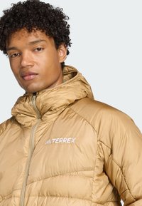 Tamna izolirana jakna s kapuljačom, sa prešanim dijelovima i prednjim patent zatvaračem. "TERREX" logo prikazan u bijeloj boji na prsima.