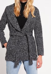 Abrigo de punto en blanco y negro, con cuello, manga larga, cinturón con lazo y tejido texturizado. Lleva puesto sobre una camiseta negra.