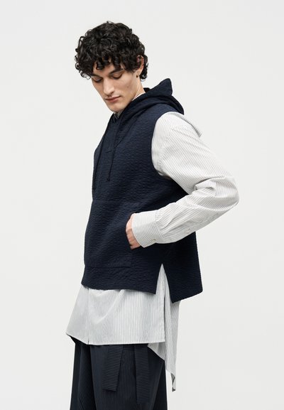 Jonge man met krullend haar, gekleed in een marineblauwe mouwloze hoodie over een gestreept shirt met lange mouwen en brede marineblauwe broek, staand in zijaanzicht.