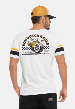Homme portant un t-shirt blanc à thème course avec un graphisme de pneu en flammes et le texte "Von Dutch Racer", pantalon noir et casquette jaune, vu de dos.