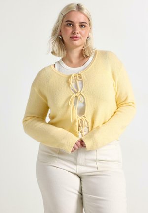 Donna con capelli biondi indossa un cardigan giallo pallido annodato con tre fiocchi sopra una maglia bianca e pantaloni a vita alta color crema, in piedi su uno sfondo bianco.