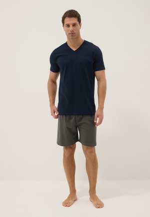 Hombre de pie descalzo sobre fondo liso con camiseta azul marino de cuello en V y pantalones cortos gris oscuro.