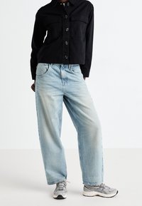 Mustan napilla varustettu takki, jossa on rintataskut, yhdistettynä vaaleansinisiin leveisiin farkkuihin. Harmaat lenkkarit täydentävät asua. Minimalistinen muotoilu.