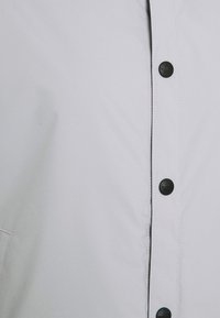 Chaqueta gris claro con un acabado mate suave, cierre de botones en la parte delantera y botones negros con detalles de logo. Tela texturizada.
