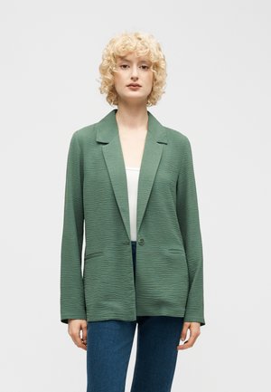 Ung kvinne med kort, krøllete blondt hår iført en teksturert grønn blazer over en hvit topp og blå jeans, stående foran en ensfarget bakgrunn.