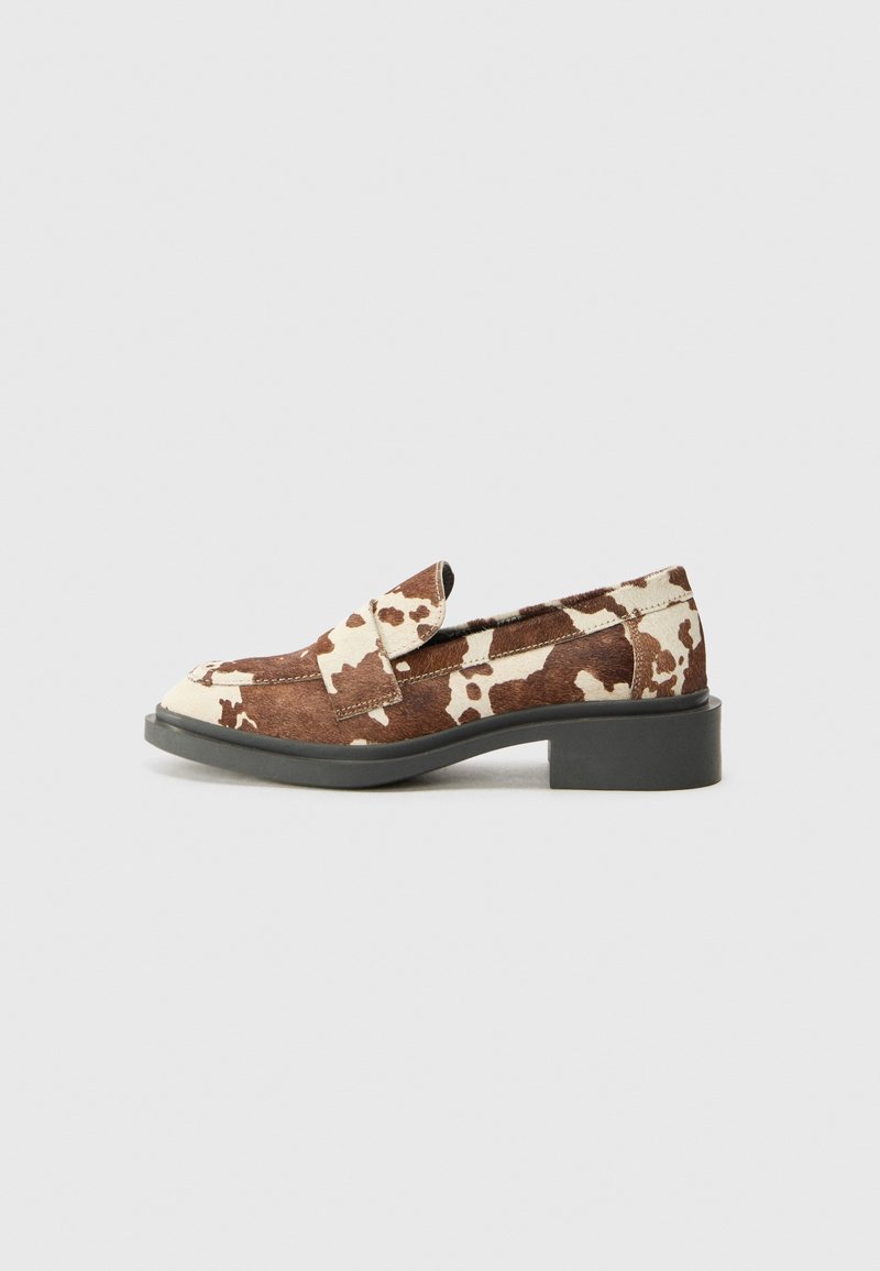 Mocassin imprimé vache marron et blanc avec semelle noire et petit talon bloc, vue de côté sur fond gris clair.