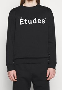 Sudadera negra de algodón, con cuello redondo y puños acanalados, y con un llamativo texto en blanco "Études" en la parte delantera.