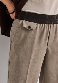 Pantalon marron clair avec une texture douce, doté d'une ceinture élastique noire et d'un bouton décoratif sur la poche avant.