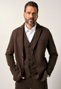 Giacca marrone con revers a punta, due tasche anteriori e chiusura con bottone, indossata sopra una camicia bianca, caratterizzata da una texture liscia.