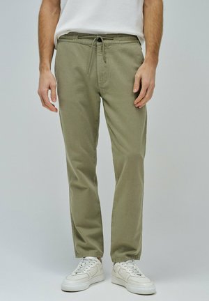 Pantalon vert olive avec une taille à cordon, au style décontracté et avec des poches latérales. Porté avec des baskets blanches.