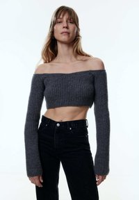 Grauer, schulterfreier, gekürzter Pullover mit gerippter Textur und langen Ärmeln, kombiniert mit hoch geschnittenen schwarzen Jeans mit Knopfverschluss.