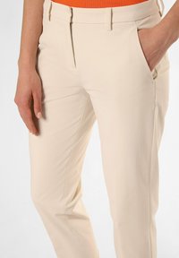 Beige Hose mit schlankem Schnitt, ausgestattet mit Seitentaschen und einem glatten, leichten Stoff. Der Bund ist teilweise sichtbar und wird mit einem orangefarbenen Oberteil kombiniert.