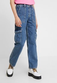 Pantalones cargo de mezclilla azul con un ajuste holgado, que cuentan con múltiples bolsillos laterales y un diseño de cintura alta, combinados con botas blancas de plataforma.