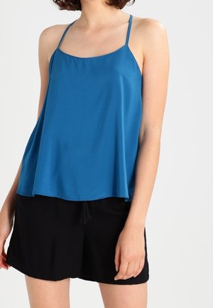 Nainen, jolla on päällään löysä sininen spagettihihnallinen toppi ja mustat shortsit, seisoo yksiväristä vaaleaa taustaa vasten.