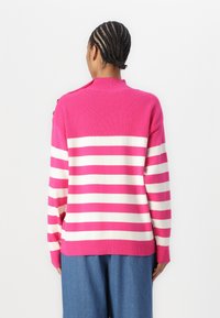 Pull en maille rayé avec des rayures alternées roses et blanches, présentant un col montant et des poignets côtelés. Le pull est ample.