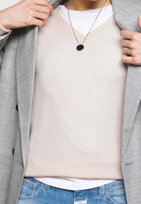 Personne portant un pull à col en V rose clair sur une chemise blanche, un collier pendentif argenté, un blazer gris clair et un jean bleu, ajustant le col du blazer.