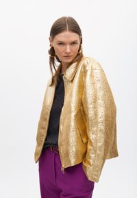 Metalni zlatni sako s reljefnom teksturom, koji ima ovratnik, zatvaranje na patentni zatvarač i bočnu džep. Uparen s crnom majicom i ljubičastim hlačama.