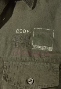 Olivgrüner Stoff mit gesticktem Text "CODE: 84", einem aufgenähten Patch und einer Knopftasche unter dem Text auf einem Kleidungsstück.