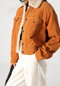 Giacca in velluto a coste arancione con colletto in shearling beige, chiusura con bottoni e due tasche sul petto. Il modello indossa pantaloni color crema e tiene in mano una borsa nera.