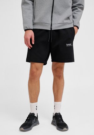 Mężczyzna ubrany w czarne spodenki Hummel, szarą kurtkę zapinaną na zamek, białe skarpetki z czarnymi szewronami oraz czarne buty sportowe, stoi z rozluźnionymi rękami.