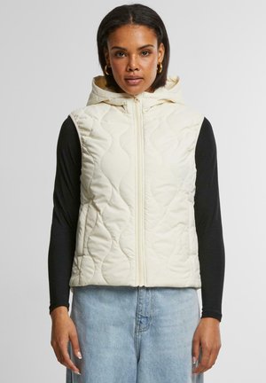 Gepolsterte cremefarbene Weste mit Kapuze und Frontreißverschluss, die ein schlankes Design und ein strukturiertes Muster aufweist, über einem schwarzen Langarmshirt getragen.