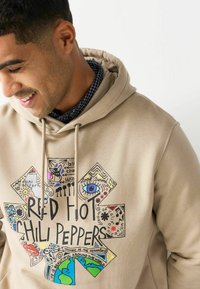Beige hoodie med et farverigt grafisk design, der inkluderer forskellige mønstre og tekst, herunder "RED HOT CHILI PEPPERS" tydeligt vist.