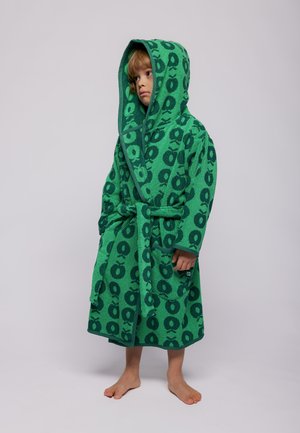 Barn iført en grøn hættetrøje badekåbe med mørkegrønne æblemønstre, stående barfodet mod en ensartet lys baggrund.