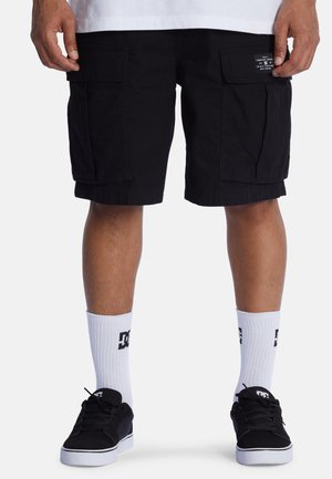 Schwarze Cargo-Shorts mit großen Seitentaschen, aus Baumwollstoff. Getragen mit weißen Socken mit Logo und schwarzen Sneakers.