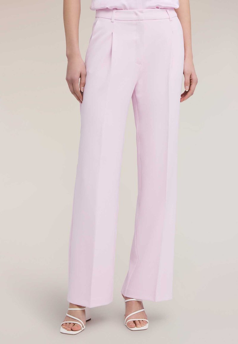 Femme portant un pantalon taille haute à jambes larges de couleur rose pastel, associé à des sandales blanches à brides, debout sur un fond neutre.