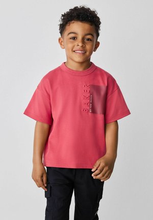 Jeune garçon aux cheveux courts et bouclés souriant, portant une chemise rouge à manches courtes avec une poche poitrine et un pantalon cargo noir.