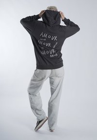 Sweat à capuche noir avec le texte « AMOUR » en blanc de tailles et orientations variées dans le dos. Assorti à un pantalon gris clair et des baskets noires.