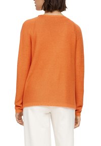 Oranger Strickpullover mit gerippter Struktur, Rundhalsausschnitt und langen Ärmeln. Kombiniert mit hellfarbigen, locker sitzenden Hosen.