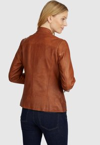 Braune Lederjacke mit taillierter Passform, minimalen Nähdetails und Stehkragen. Kombiniert mit dunkelblauen Jeans.