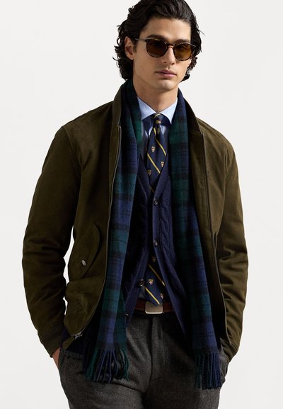 Veste en daim vert olive sur un pull en laine bleu marine à mailles torsadées, chemise bleue et cravate à motifs ; pantalon en laine gris ; écharpe à carreaux avec franges.