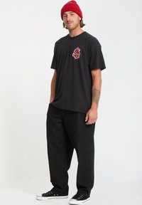 Czarny t-shirt z kolorowym nadrukiem na klatce piersiowej, zestawiony z luźnymi czarnymi spodniami i czarnymi trampkami. Model nosi czerwoną czapkę beanie.