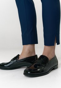 Zwarte loafers van lakleer met een lage hak, voorzien van een ronde neus en een decoratieve band over de wreef, gecombineerd met marineblauwe broek.