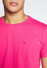 Camiseta de algodón rosa con cuello redondo, que presenta un pequeño logo bordado en el lado izquierdo del pecho en colores rojo, blanco y azul.