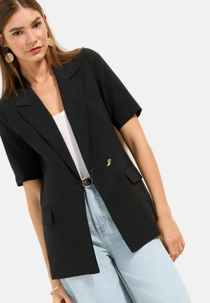 Nő fekete rövid ujjú blézert visel fehér felső fölött, világoskék farmert, kerek övcsatot és nagy kerek fülbevalókat, fehér háttér előtt.