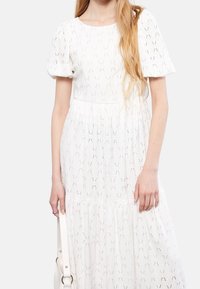 Kobieta w białej sukience z puchem i bufkami trzyma białą torebkę przy boku na tle jednolitym.