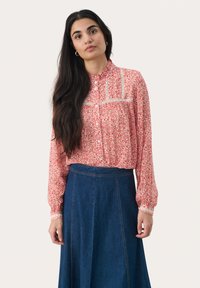 Blusa floreale rossa con maniche lunghe e chiusura a bottoni, con occasione in pizzo sul colletto. Abbinata a una gonna di jeans blu.