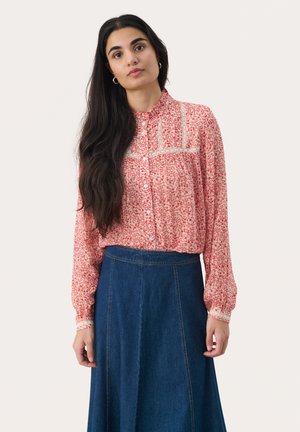 Blusa floreale rossa con maniche lunghe e chiusura a bottoni, con occasione in pizzo sul colletto. Abbinata a una gonna di jeans blu.