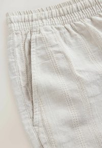 Pantalon en tissu beige clair avec une ceinture élastique et des rayures texturées verticales, avec une poche avant sur le côté gauche.
