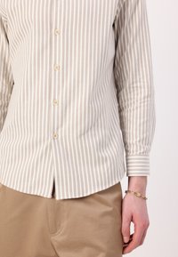 Beige og hvit stripete skjorte med lange ermer, med avrundet kant og lett tekstur. Kombinert med beige chinos og gullarmbånd.