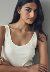 Débardeur blanc côtelé avec un col en U, présentant une coupe ajustée. Le mannequin a de longs cheveux noirs et est posé sur un fond gris.
