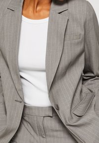 Blazer de rayas gris hecho de una tela de textura suave. Cuenta con un solo botón, solapas de muesca y un bolsillo en el pecho. Se lleva sobre una blusa blanca.