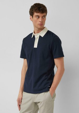 Jonge man staand met handen in zijn zakken, gekleed in een marineblauw poloshirt met een witte kraag en lichtbeige broek, kijkend naar links.