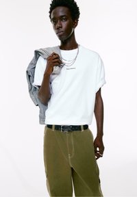 Ung mand med mørk hud iført en hvid t-shirt med teksten "Simple Moments", olivengrønne bukser, mens han holder en jeansjakke over den ene skulder.
