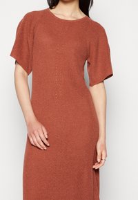 Rustfarbenes Strickkleid mit kurzen Ärmeln, das ein strukturiertes Rippmuster und vertikale Reihen kleiner Öffnungen an der Vorderseite aufweist.